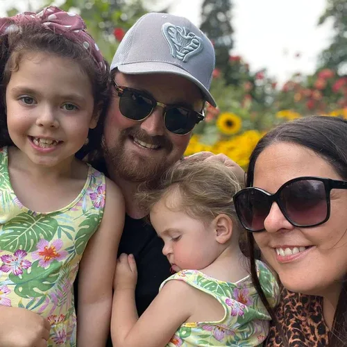 A family of four poses outdoors, surrounded by sunflowers. The two children wear matching floral dresses, and the adults wear sunglasses.