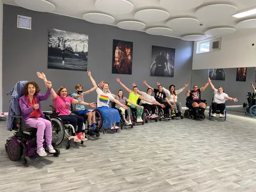A group of people in wheelchairs joyfully raise their arms in a dance studio, surrounded by wall art and mirrors.